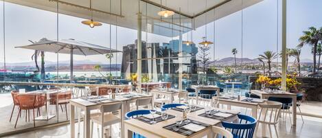 A modern restaurant with large windows offering a view of the city and mountains. Tables are neatly arranged with blue and white chairs.