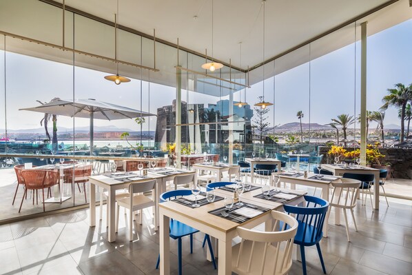 A modern restaurant with large windows offering a view of the city and mountains. Tables are neatly arranged with blue and white chairs. - Barceló Fuerteventura Castillo (Antigua)