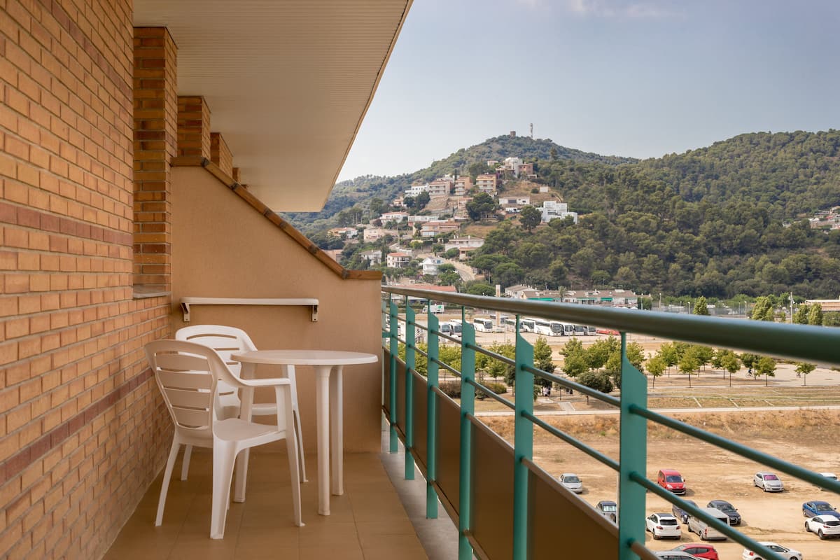 Un balcon avec des chaises en plastique blanches et une petite table, donnant sur un parking et une colline parsemée de bâtiments.
