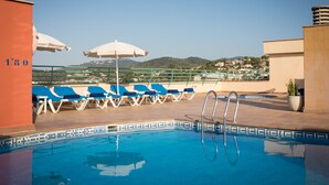 Área de piscina no terraço com espreguiçadeiras azuis e guarda-sóis brancos, com vista clara para o horizonte da cidade.