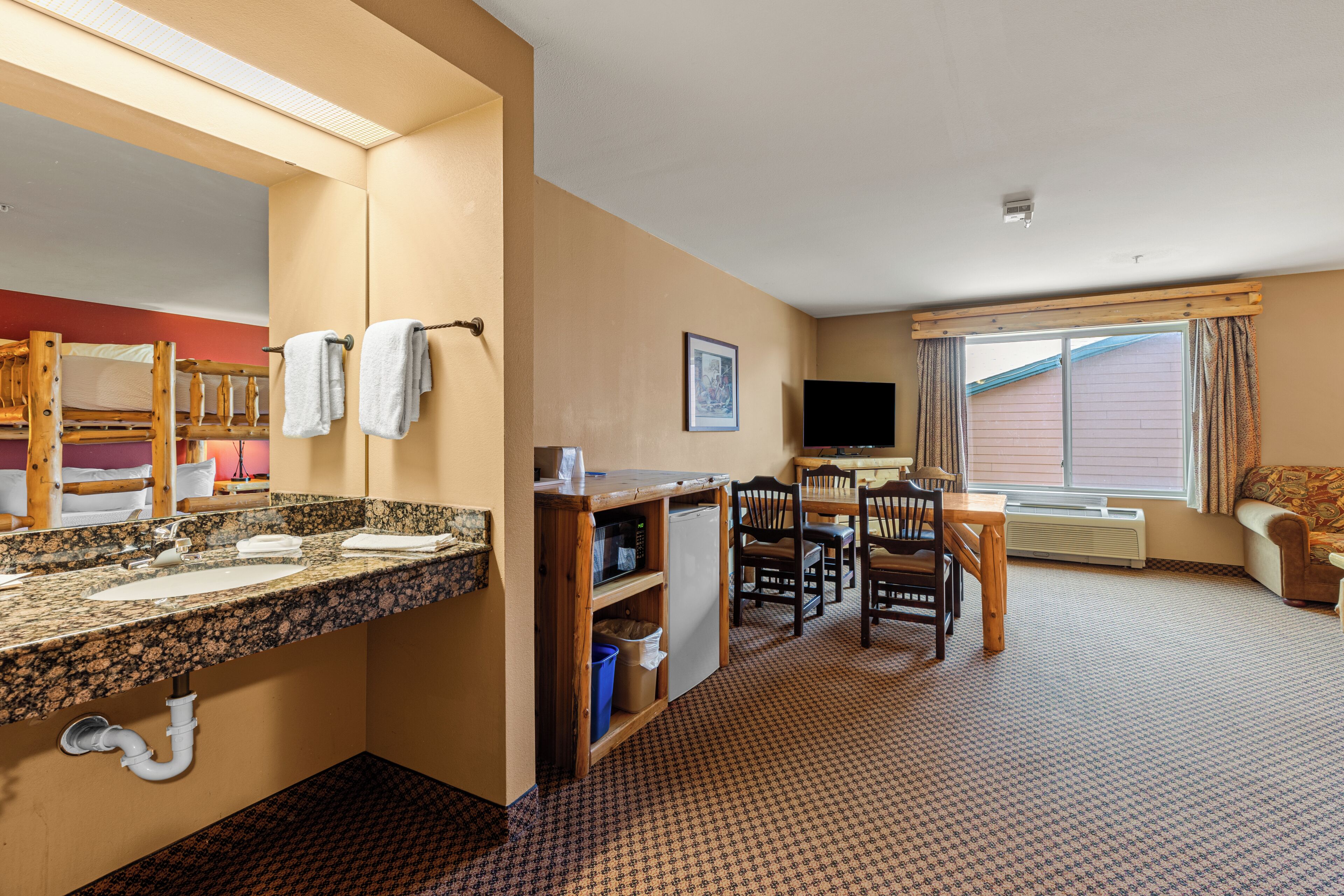 A hotel room with a bathroom, a dining area with chairs and a table, a television, and a window with curtains.