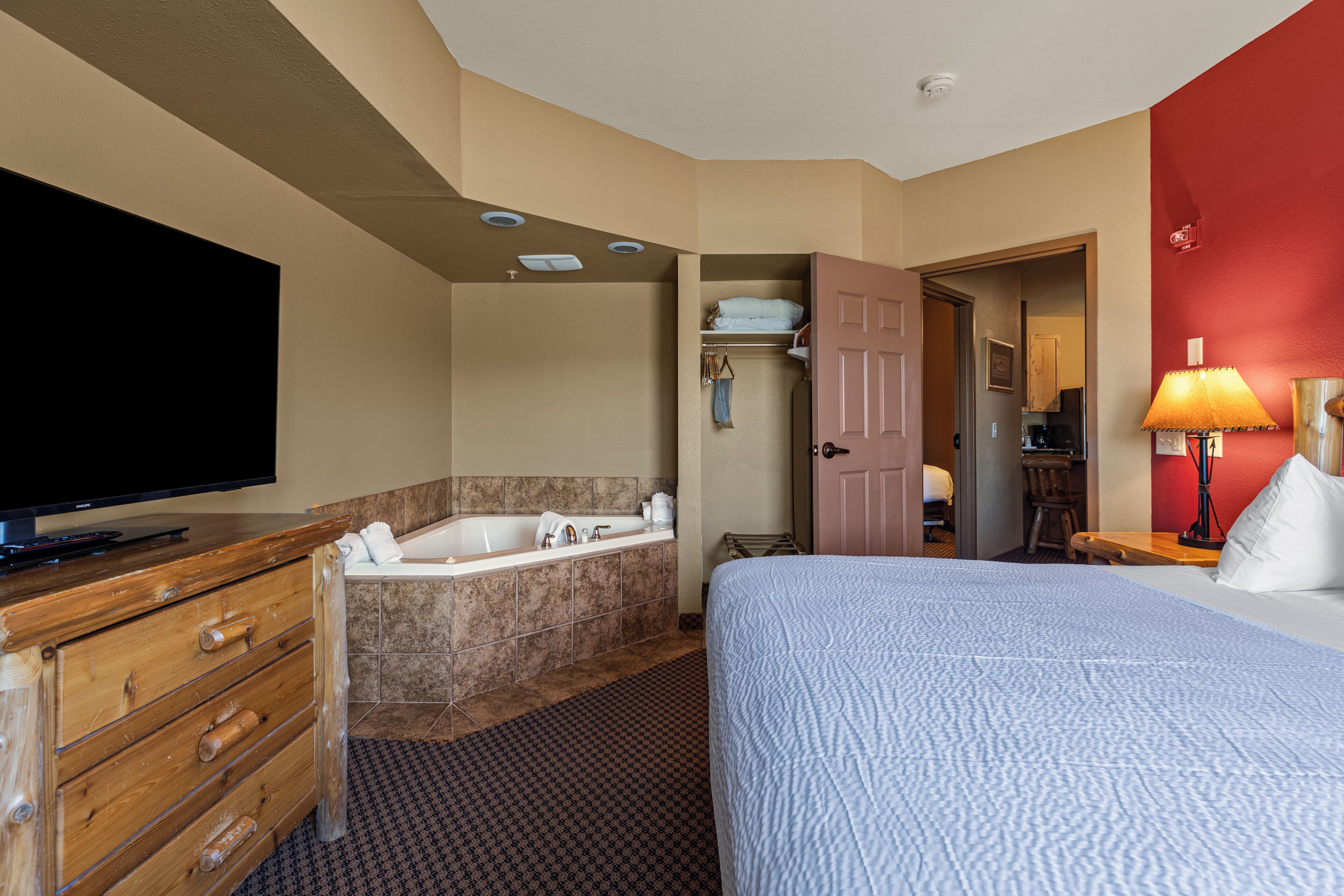 Une chambre d’hôtel comprenant un lit, une commode en bois, une télévision, une salle de bain avec baignoire et un mur d’accent de couleur rouge.