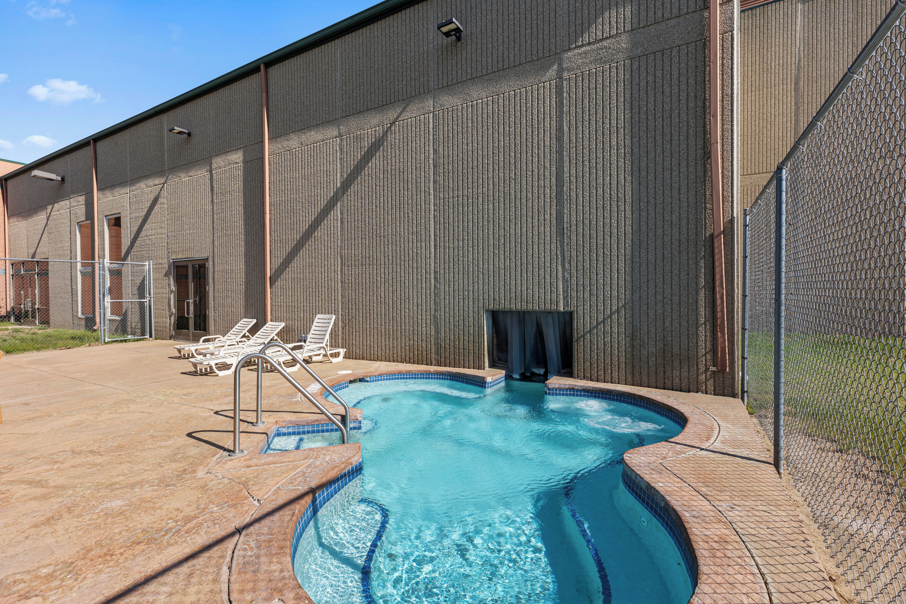 A swimming pool with a tiled edge, surrounded by a fence, and adjacent to a building with a metal exterior.