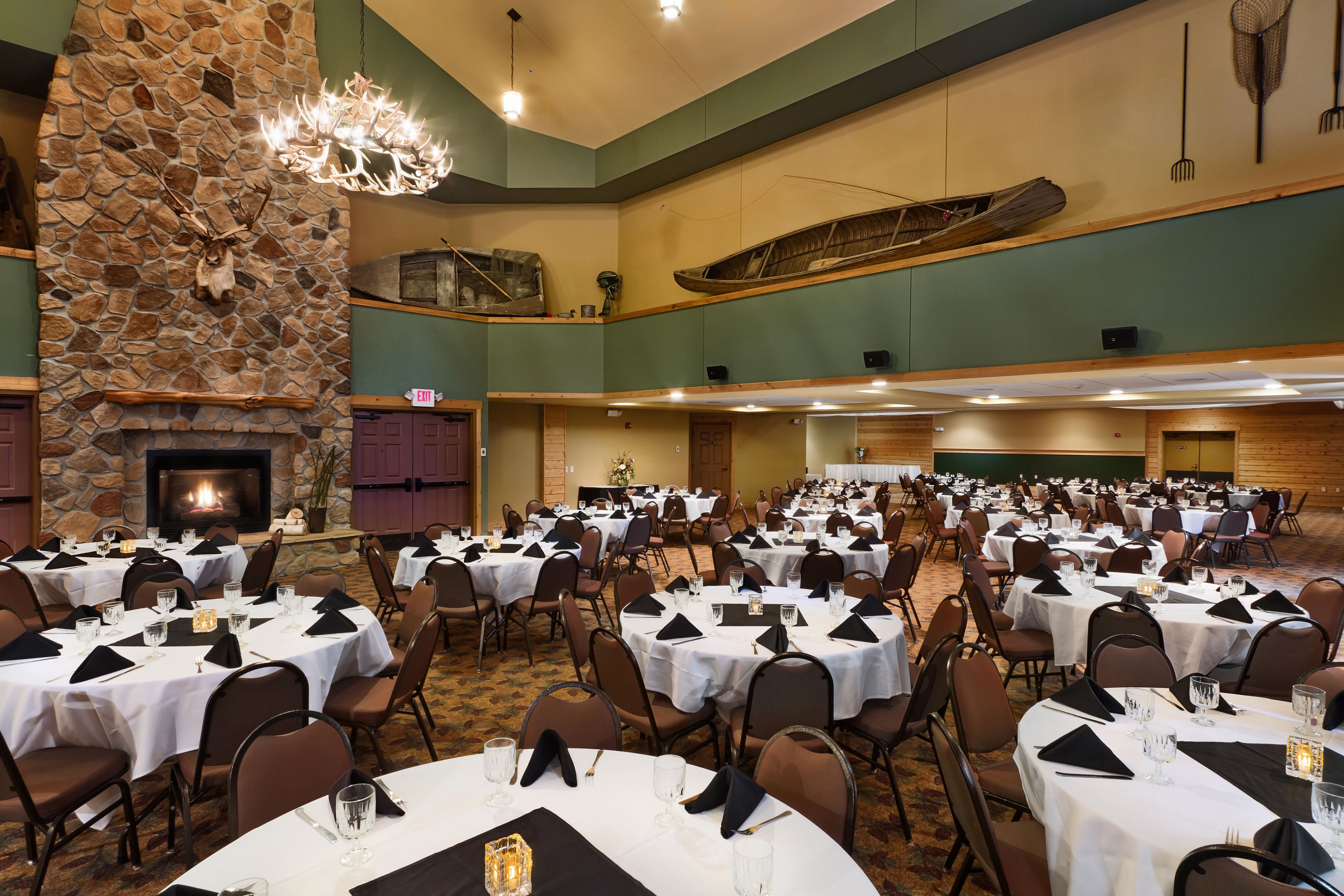 A banquet hall with multiple round tables set for dining, a stone fireplace, and a large chandelier.