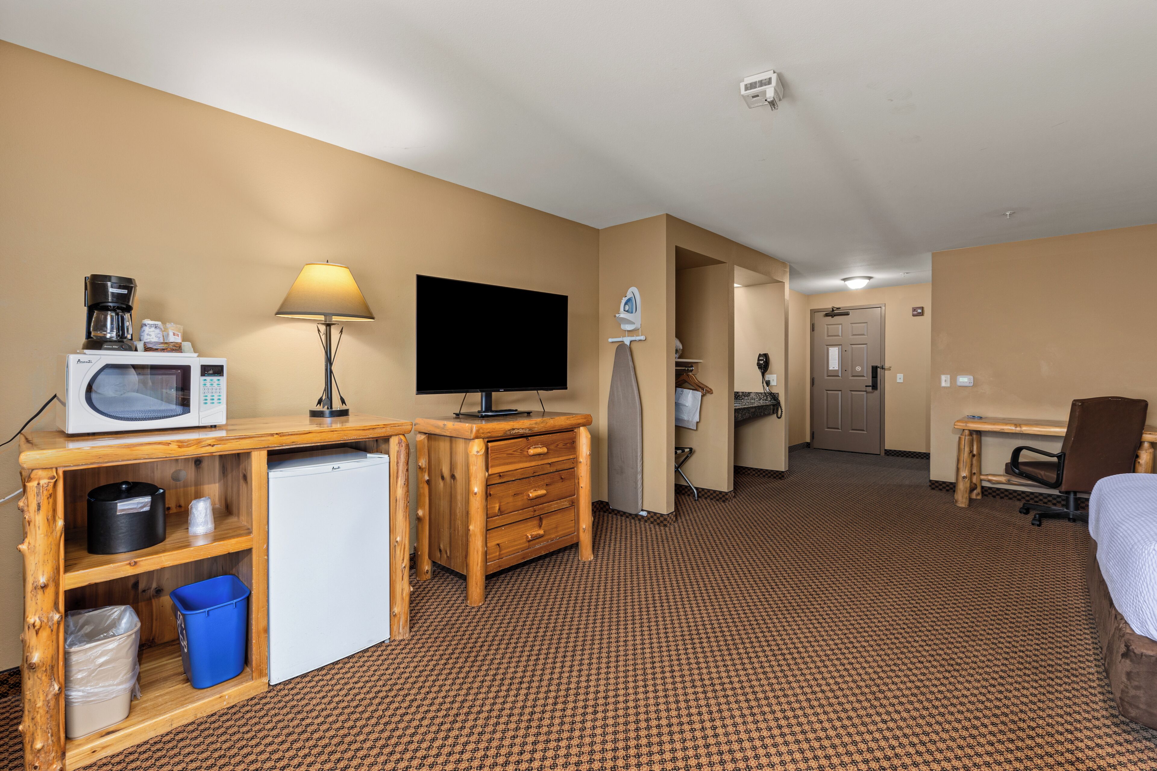 Habitación de hotel con escritorio de madera, televisor, lámpara, cama, refrigerador pequeño y un basurero.