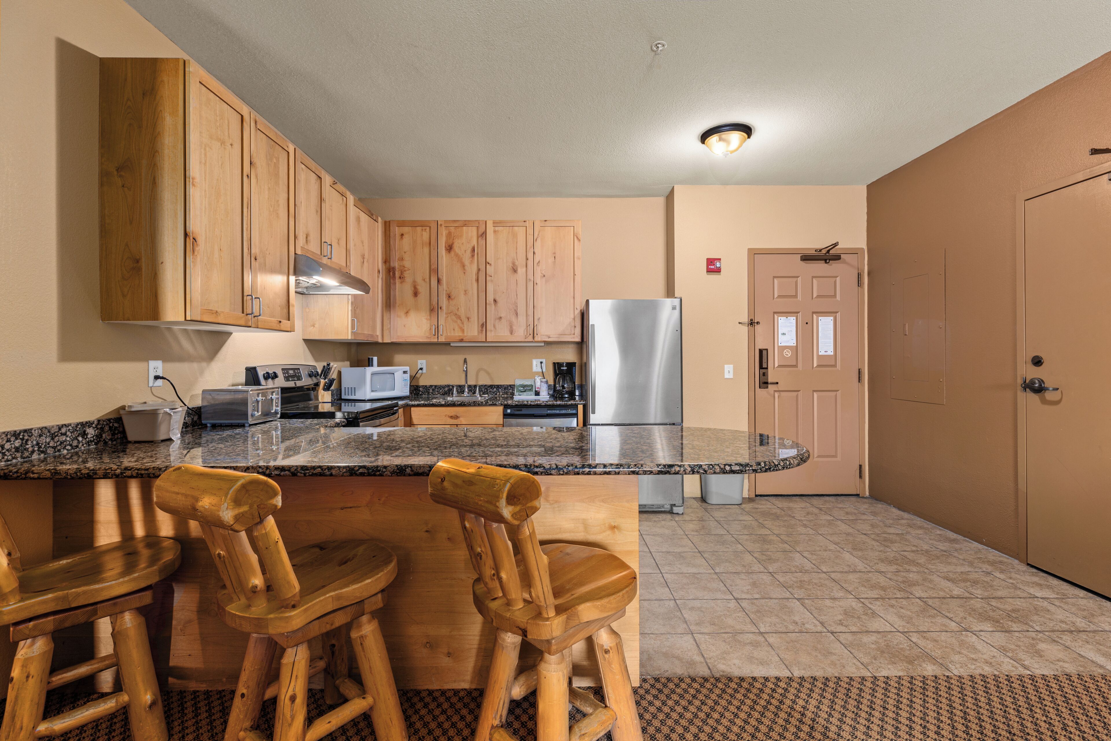A kitchen with wooden cabinets, a stainless steel refrigerator, a microwave, and a granite countertop. There are wooden bar stools and a small island with a sink.