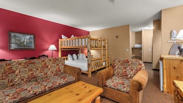 Habitación de hotel con literas, dos sofás estampados, una mesa de madera y una pared roja con un cuadro enmarcado.