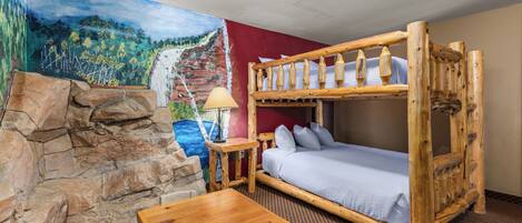 A bunk bed with a wooden frame, a small table, a lamp, and a stone wall.