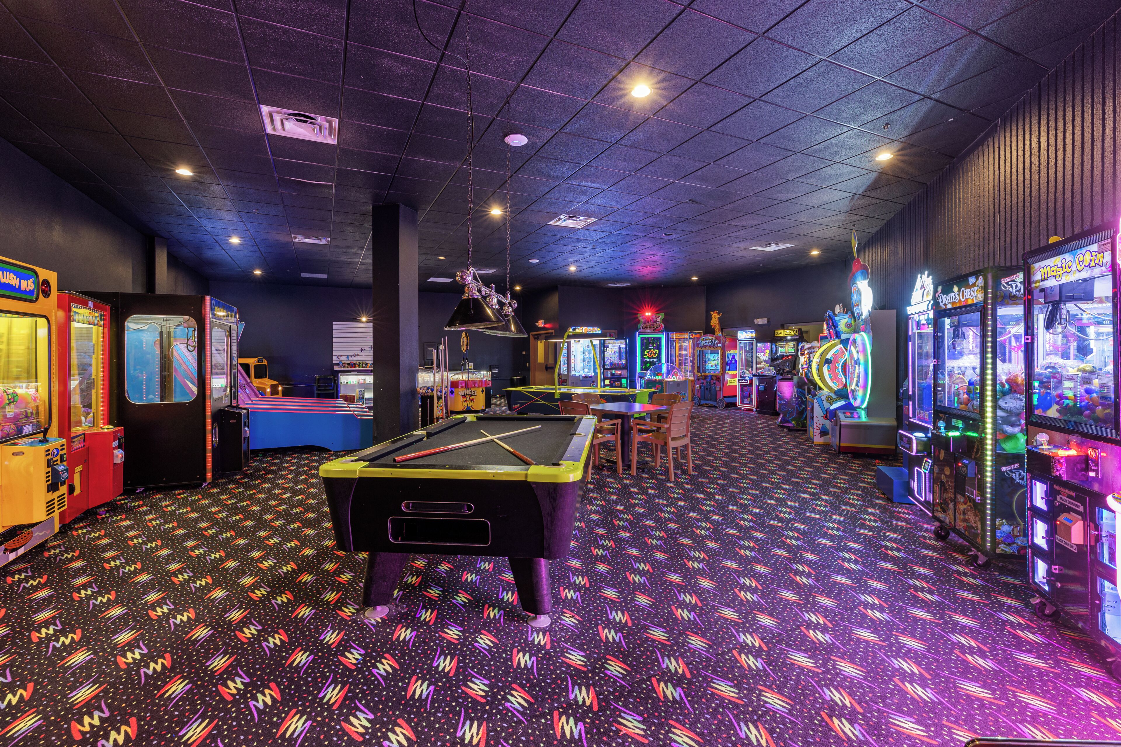 Una mesa de billar en el centro, rodeada de máquinas de arcade.