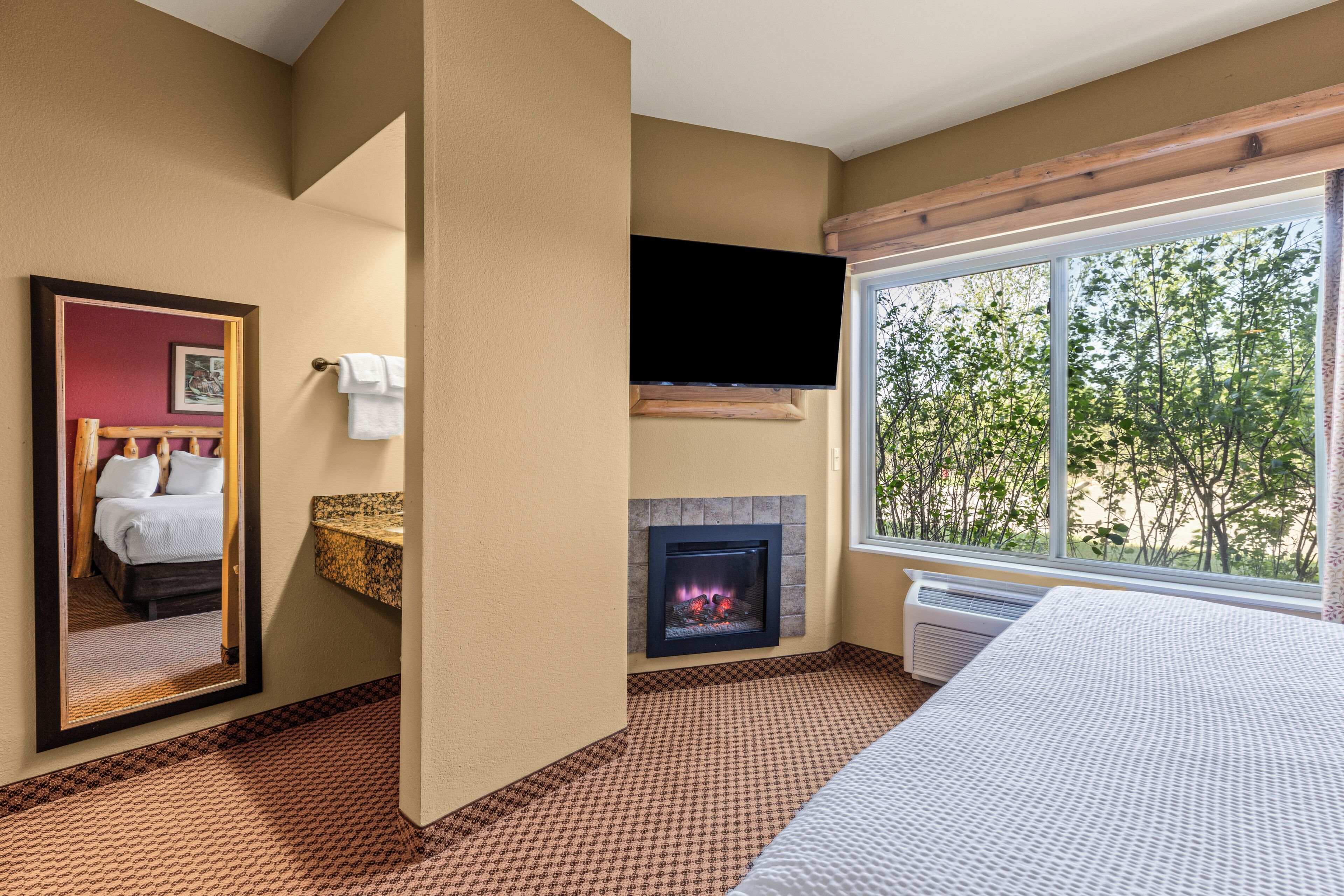 A hotel room with a bed, a fireplace, a TV mounted on the wall, and a large window offering a view of trees.
