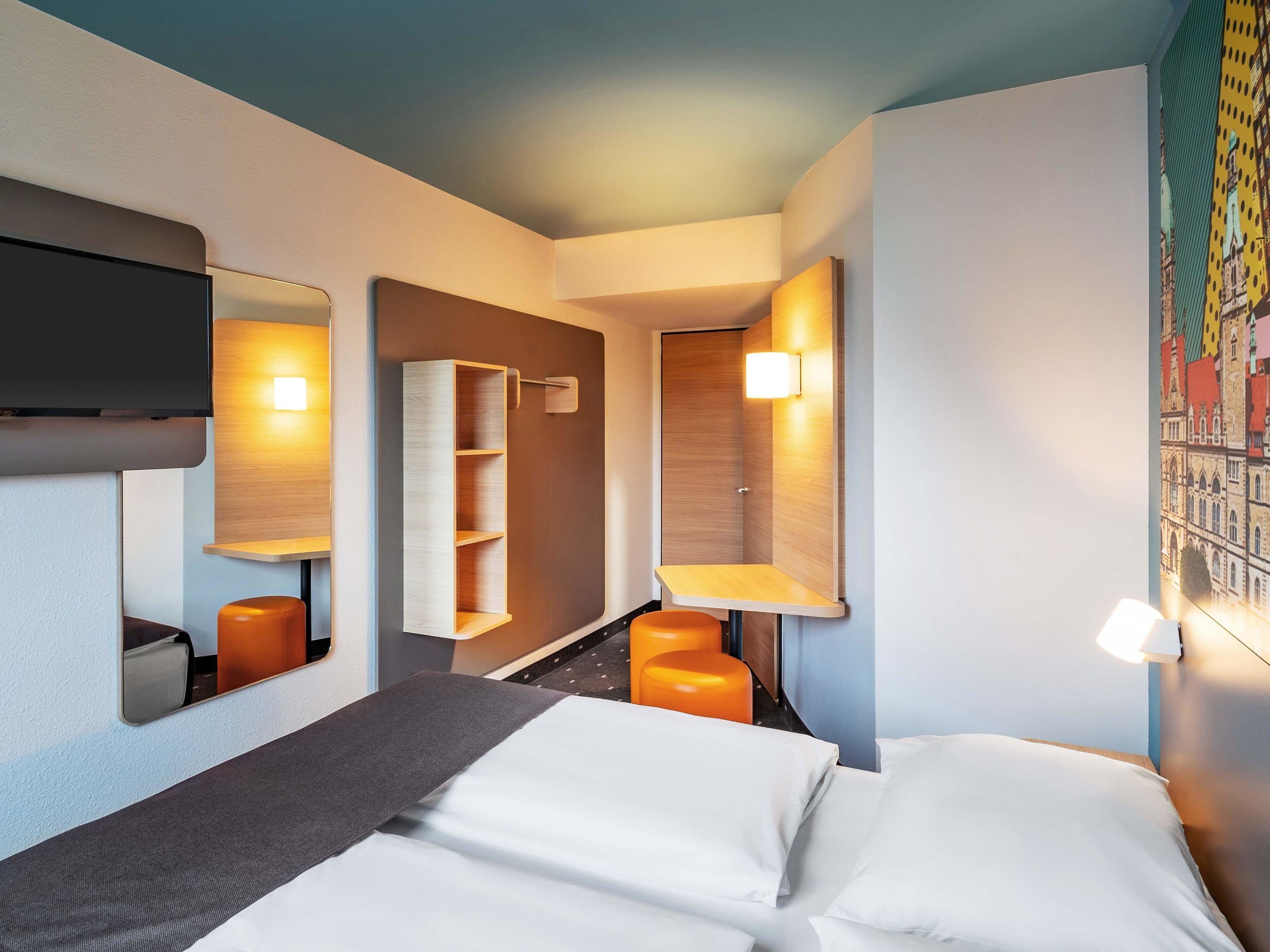 Una habitación de hotel con una cama, un escritorio, una silla, un estante y un televisor montado en la pared.