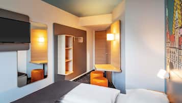 Una habitación de hotel con una cama, un escritorio, una silla, un estante y un televisor montado en la pared.