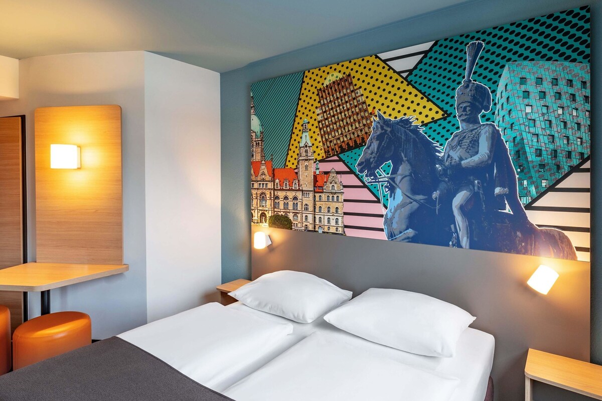 A hotel room with a bed, two pillows, a nightstand, a lamp, and a wall mural.