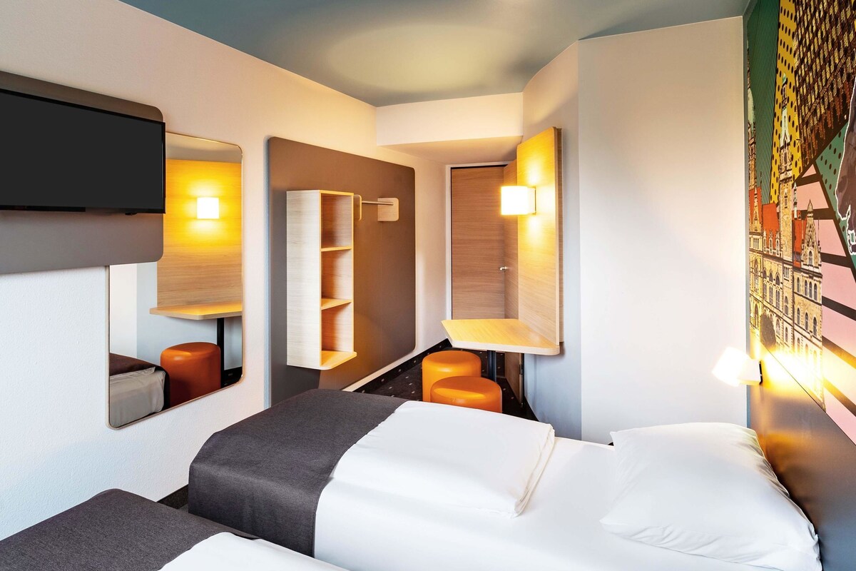A modern hotel room with a bed, a wardrobe, a mirror, and a wall-mounted shelf.