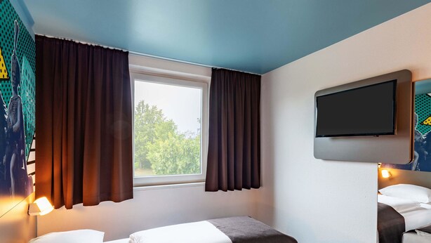 A hotel room with two beds, a TV mounted on the wall, and a window with curtains. - B&B Hotel Hannover-Lahe (Hannover)