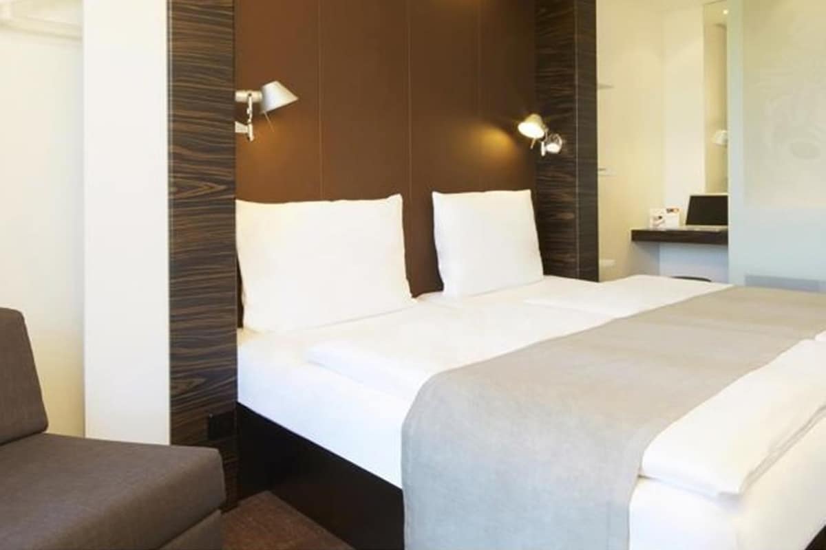 A hotel room with a large bed, two bedside lamps, a wooden headboard, and a small desk with a laptop.