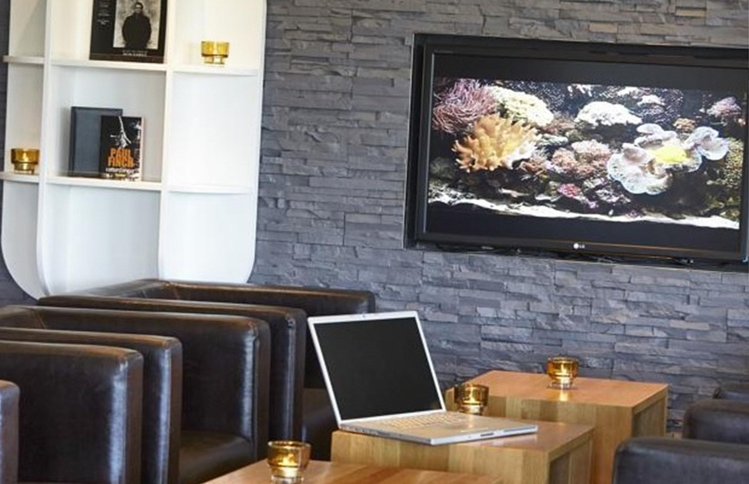 A modern lounge area with leather seating, a laptop on a wooden table, and a television mounted on a stone wall.