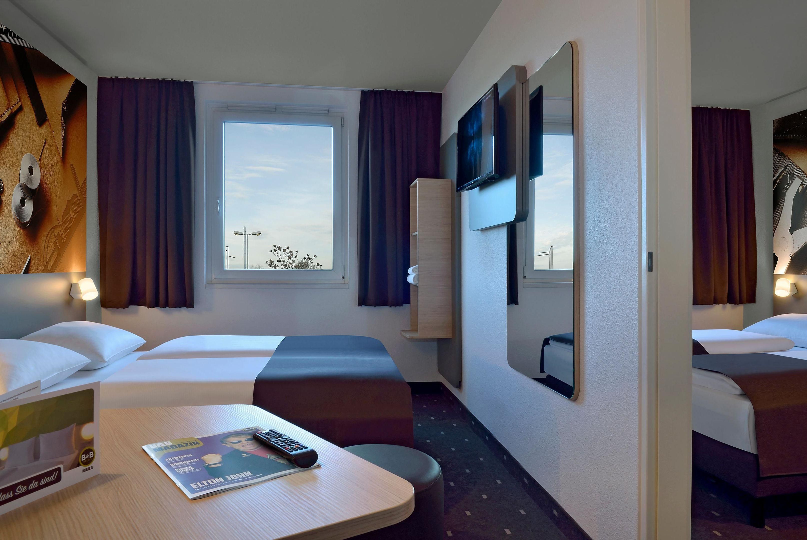 A hotel room with two beds, a window with curtains, a flat-screen TV, a table with a magazine and remote, and a mirror on the wall.
