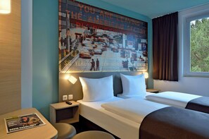 A hotel room with a large poster on the wall, a bed with white linens, a nightstand, a table with a magazine, and a window with curtains. - Premiere Classe Berlin-Dreilinden (Kleinmachnow)