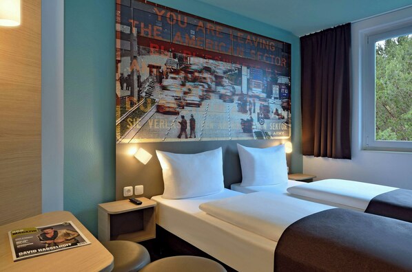 A hotel room with a large poster on the wall, a bed with white linens, a nightstand, a table with a magazine, and a window with curtains. - Premiere Classe Berlin-Dreilinden (Kleinmachnow)
