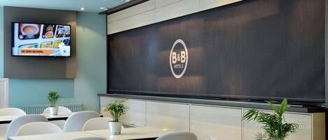 A modern hotel restaurant with a large menu display, seating area, and a B&B logo on the wall.