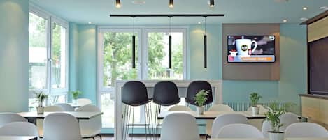 A modern dining area with white tables and chairs, large windows, and a television screen displaying a hotel advertisement.