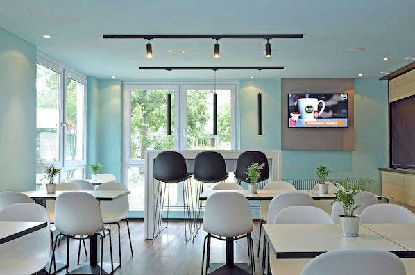 A modern dining area with white tables and chairs, large windows, and a television screen displaying a hotel advertisement.