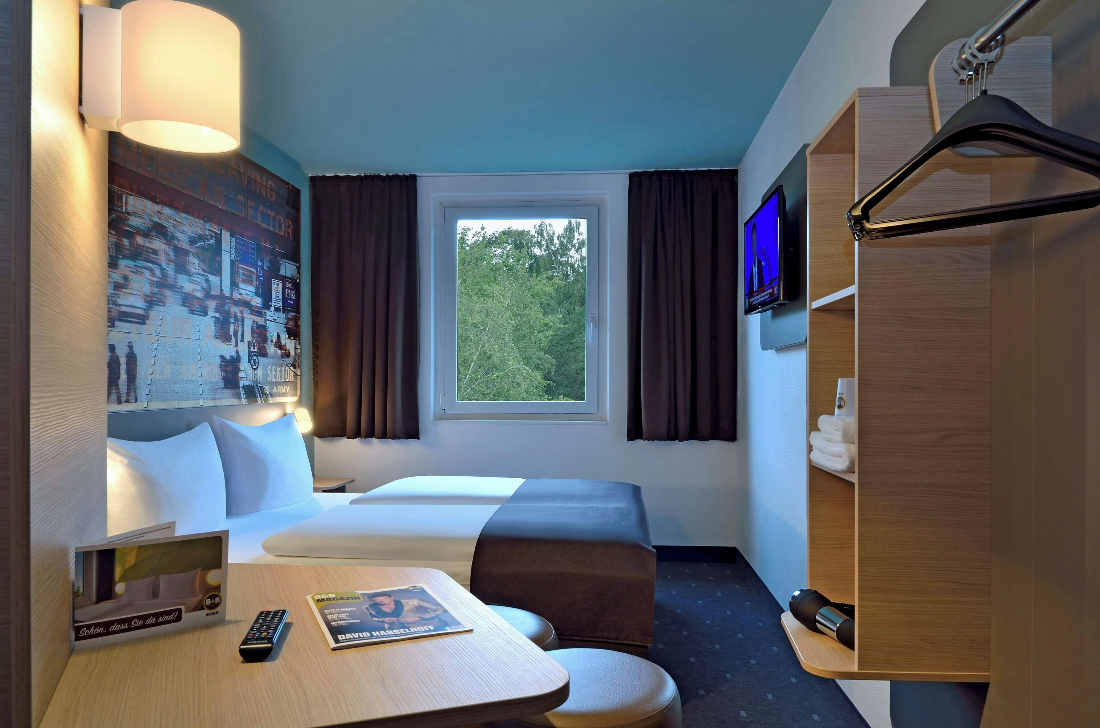 Habitación de hotel con cama, escritorio con control remoto y revista, televisor en la pared y ventana con cortinas.