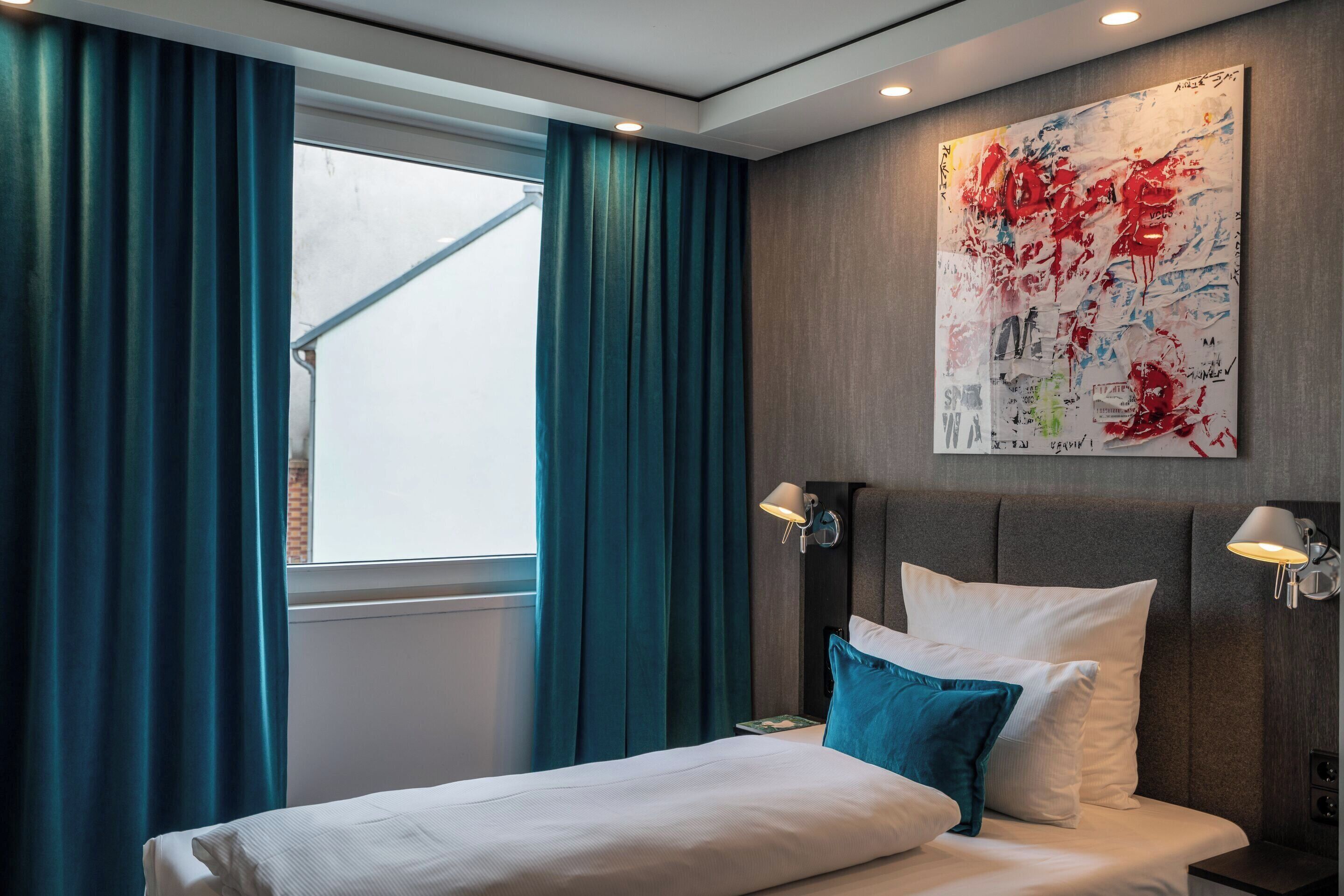 Une chambre d’hôtel moderne avec un grand lit, des rideaux de couleur turquoise, un grand tableau abstrait accroché au mur et deux lampes de chevet.