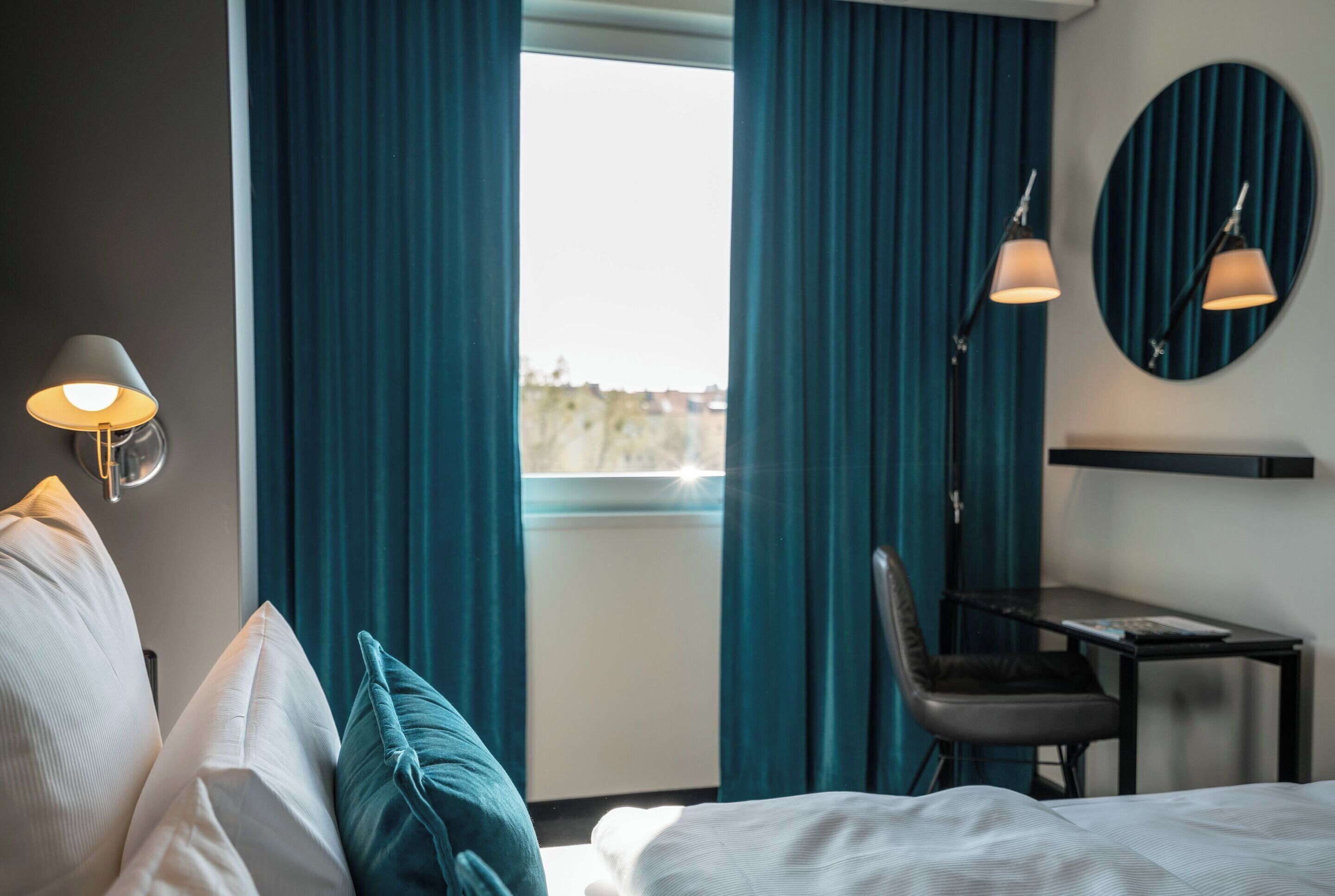 A hotel room with a bed, a desk, two lamps, and a view through a window with curtains.
