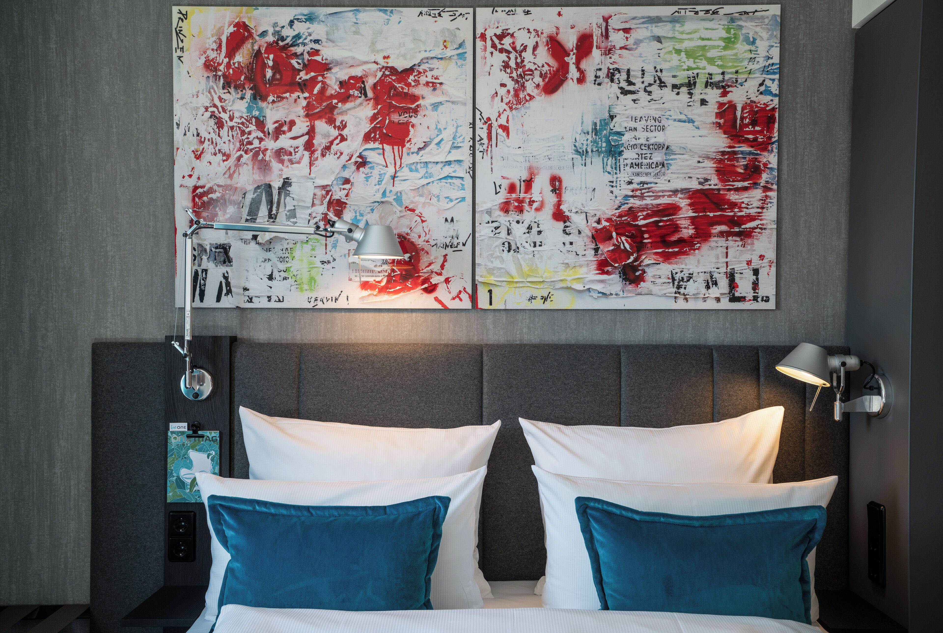 A modern hotel room with a large bed, two pillows, a nightstand, a lamp, and abstract art on the wall.