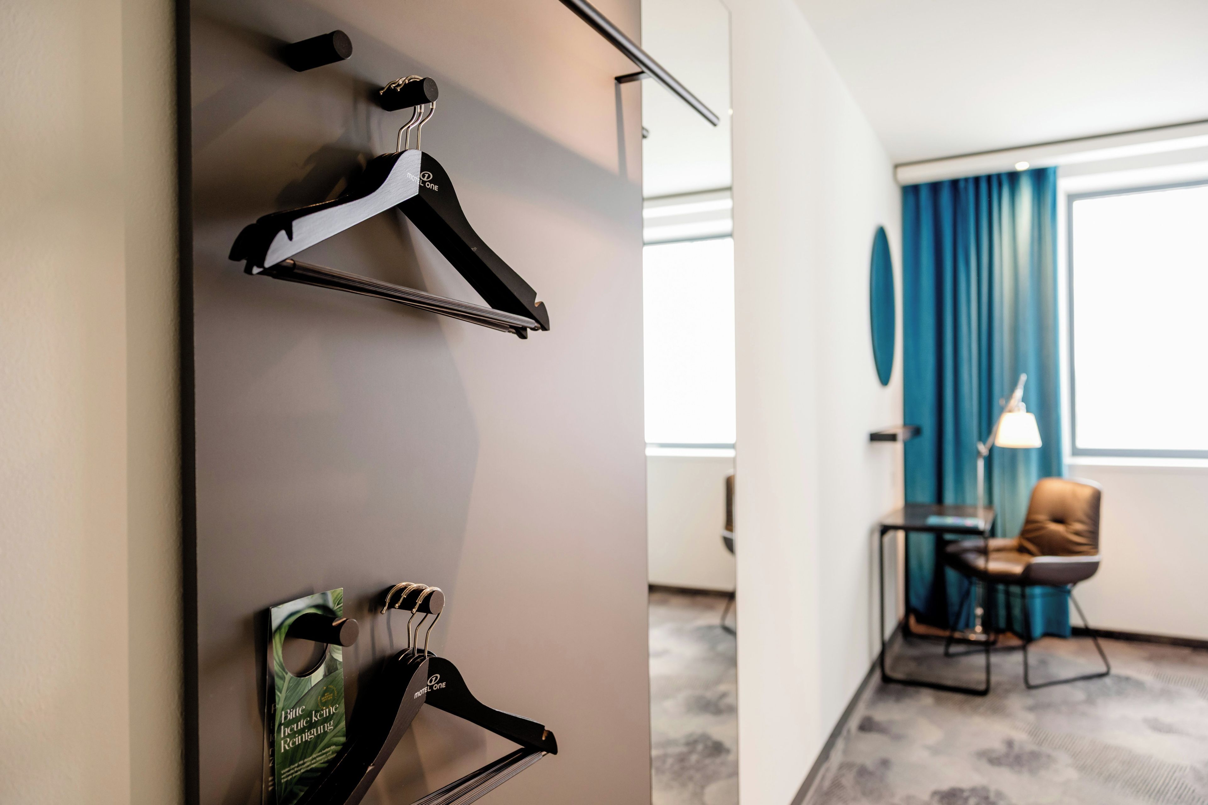 Ein Hotelzimmer mit Kleiderhaken, einem kleinen Tisch mit Lampe und Blick durch ein Fenster mit blauen Vorhängen.