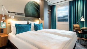 A hotel room with a large bed, a desk, a chair, a window with curtains, and a wall with a large abstract painting and a motivational quote. - Motel One Frankfurt-Airport (Frankfurt)