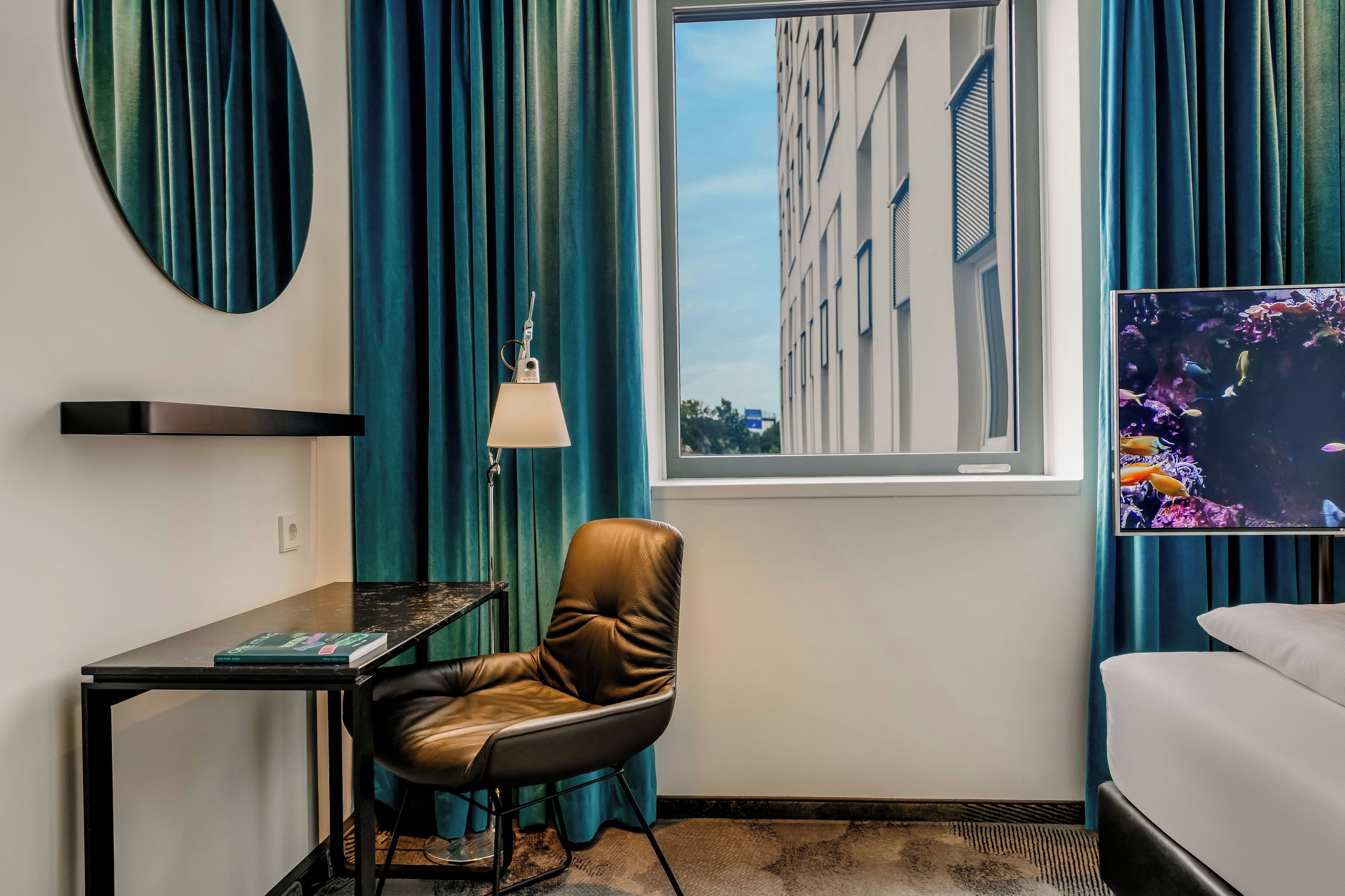 Ein Hotelzimmer mit Ledersessel, Schreibtisch mit Buch, Lampe, Bett und Fenster mit Blick auf Gebäude und ein Aquarium.