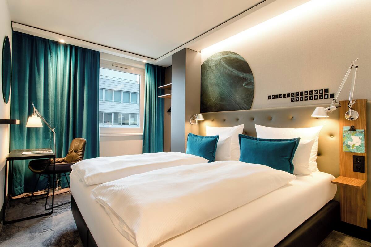 A hotel room with a large bed, a desk, a chair, and a view of a building outside.