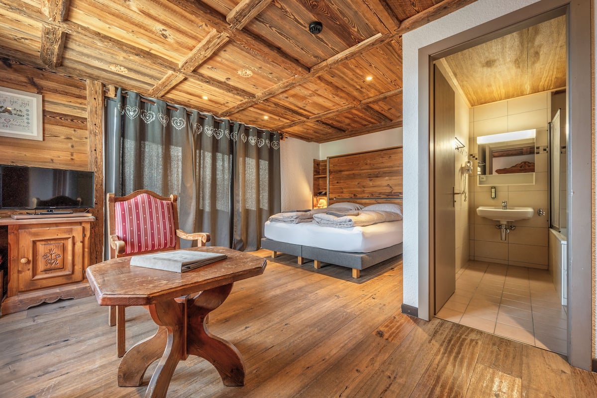 Dormitorio con piso de madera, una cama, un escritorio y un baño con lavamanos y espejo.