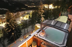 Un jacuzzi en una terraza de madera con suelo cubierto de nieve y árboles al fondo.
