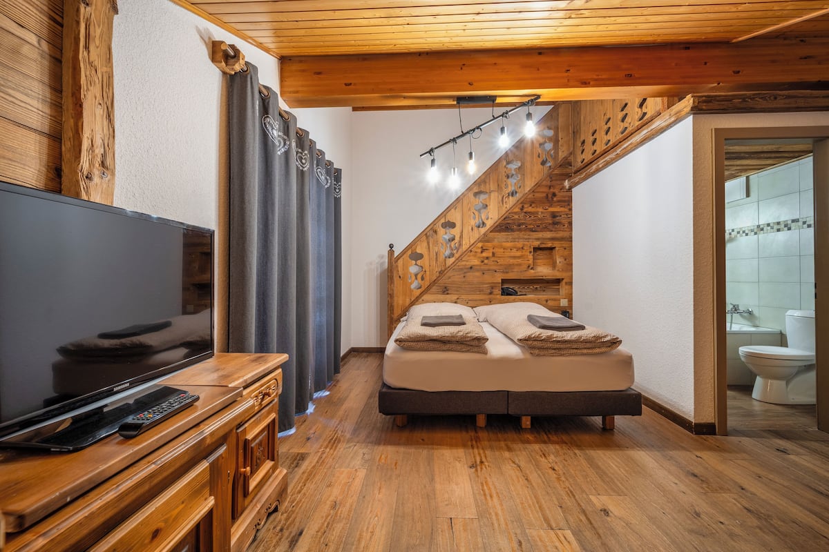 Un dormitorio con escaleras de madera, una cama con almohadas, un televisor sobre un aparador de madera y un baño visible a través de una puerta abierta.