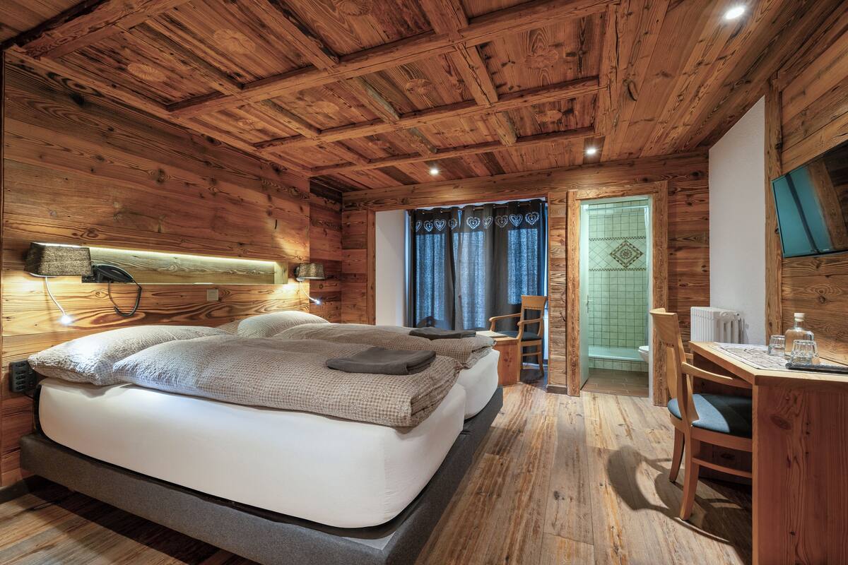 Un dormitorio tipo cabaña de madera con cama, mesita de noche, lámpara y un pequeño escritorio con silla.