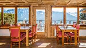 Interior de madera con mesas y sillas, ventanas amplias con vistas a la montaña y techo de madera.