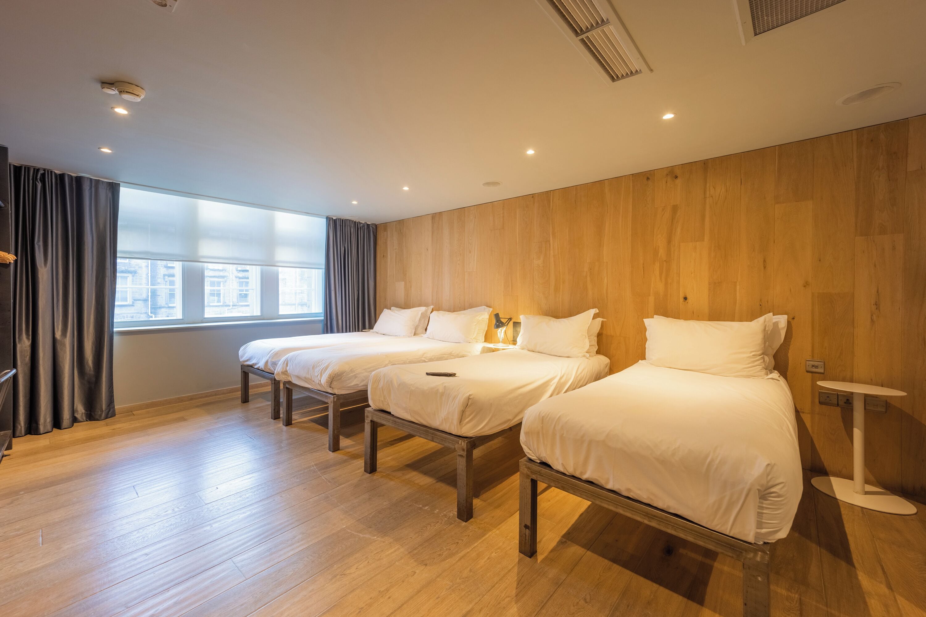 A hotel room with two beds, wooden flooring, a window with curtains, and a small bedside table.
