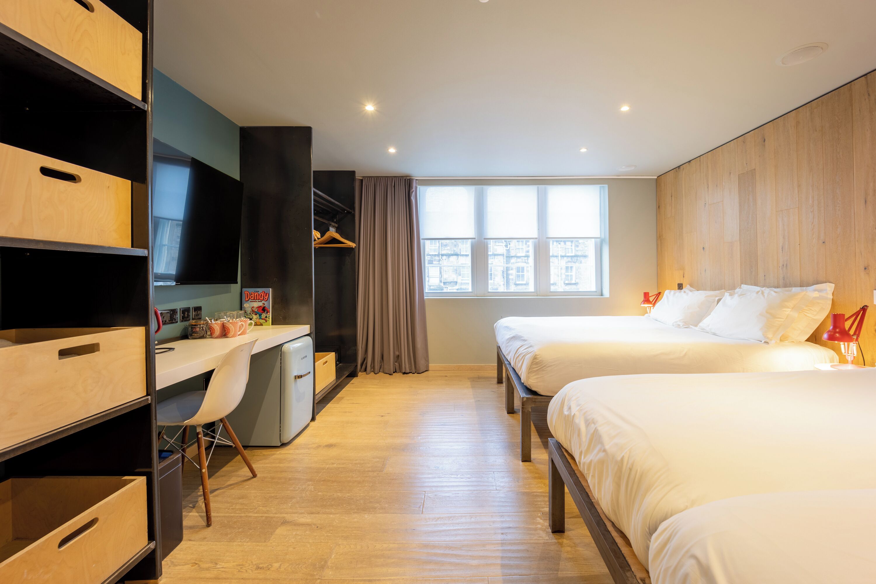 A hotel room with a desk, two beds, a wardrobe, and a window with a city view.