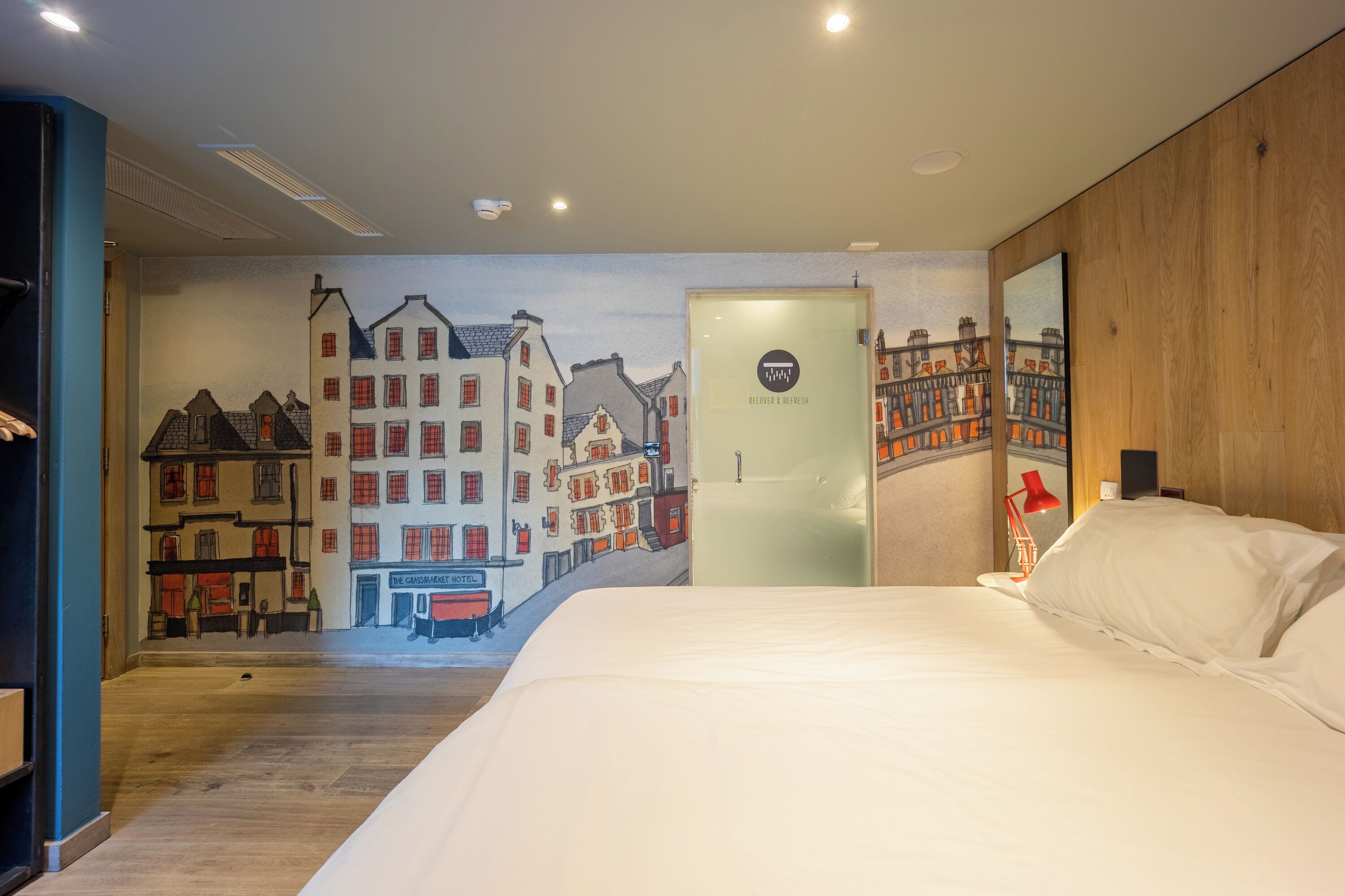Een hotelkamer met een muurschildering van een stadsgezicht, een bed met witte lakens, een rode lamp en een deur met een bordje.