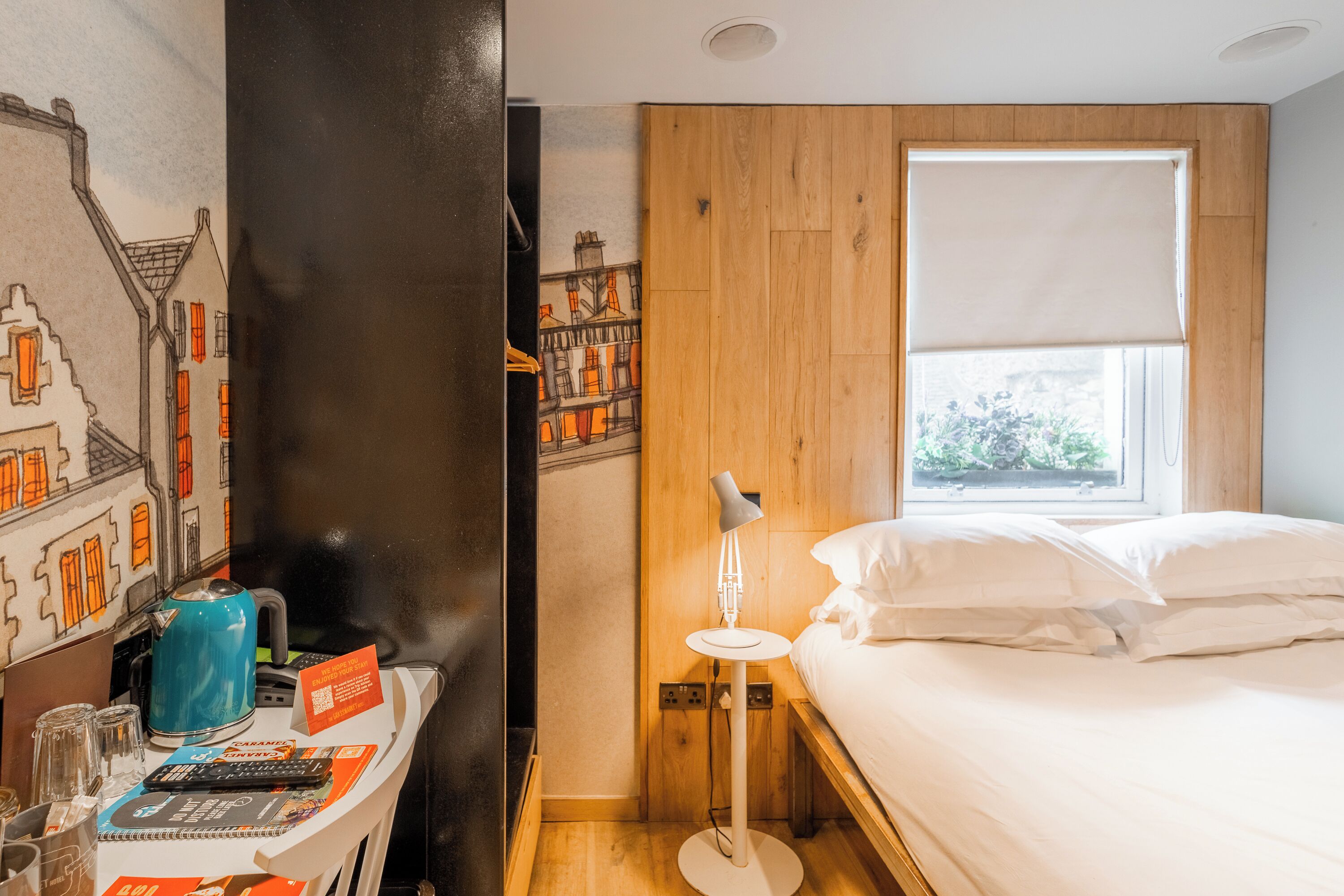 Une chambre d’hôtel avec un lit, un bureau doté d’une lampe, une fenêtre donnant sur un paysage urbain et un mur orné d’une fresque représentant une vue de la ville.