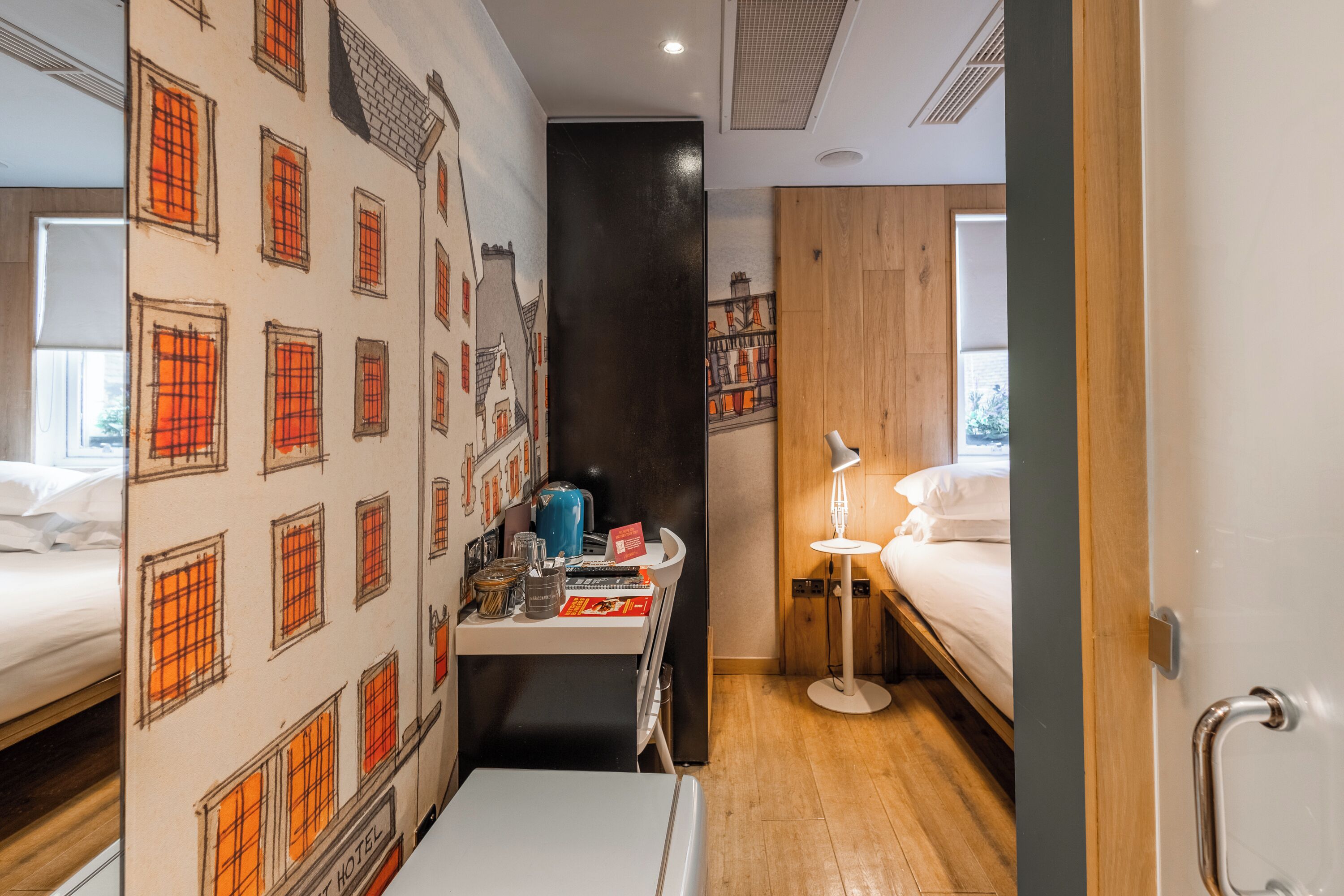 Une chambre d’hôtel avec un lit, un bureau doté d’une lampe et un mur orné d’une fresque représentant un paysage urbain.