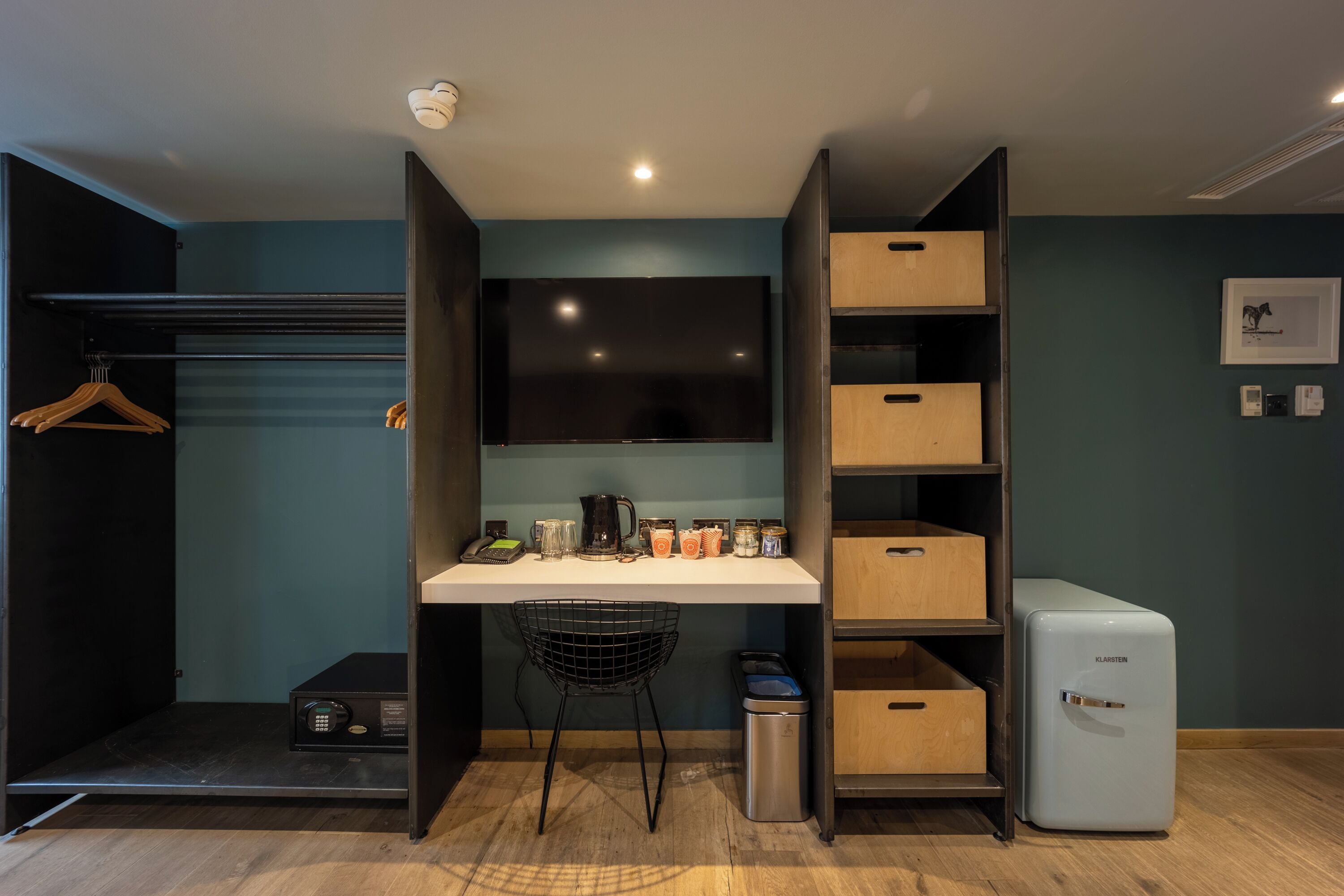 A compact room with a desk, a chair, a small trash bin, a mini-fridge, and a coat rack.
