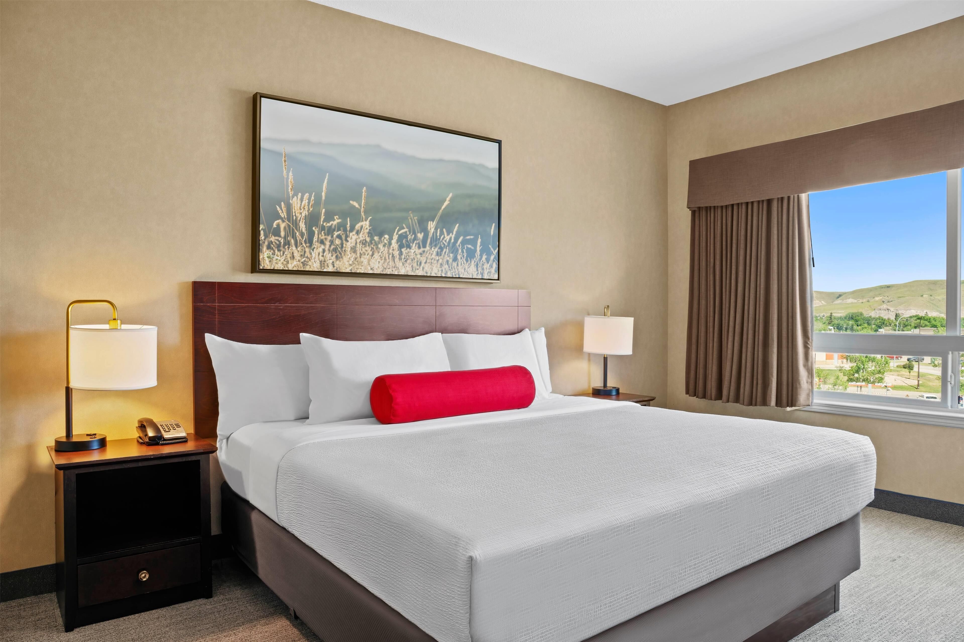 A hotel room with a large bed, two bedside tables with lamps, a painting above the bed, and a window with curtains.