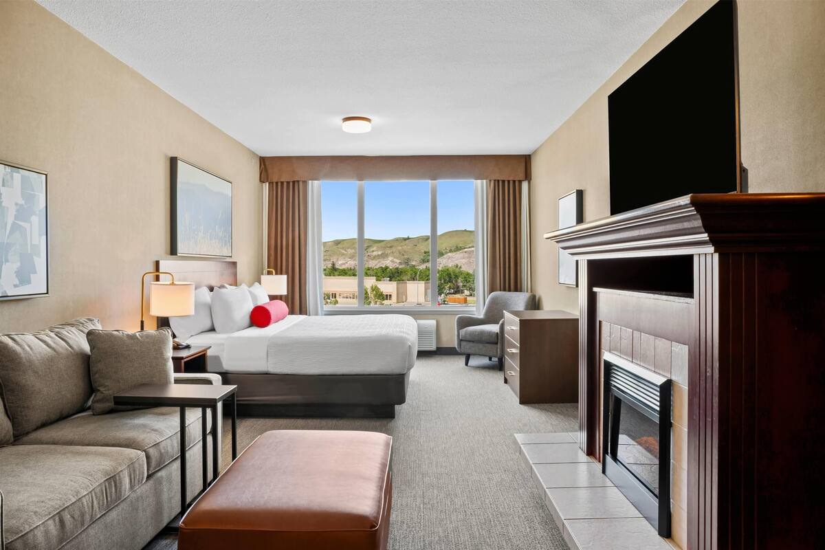 Habitación de hotel con cama, sofá, escritorio, sillón, chimenea y un ventanal con vistas a las colinas.