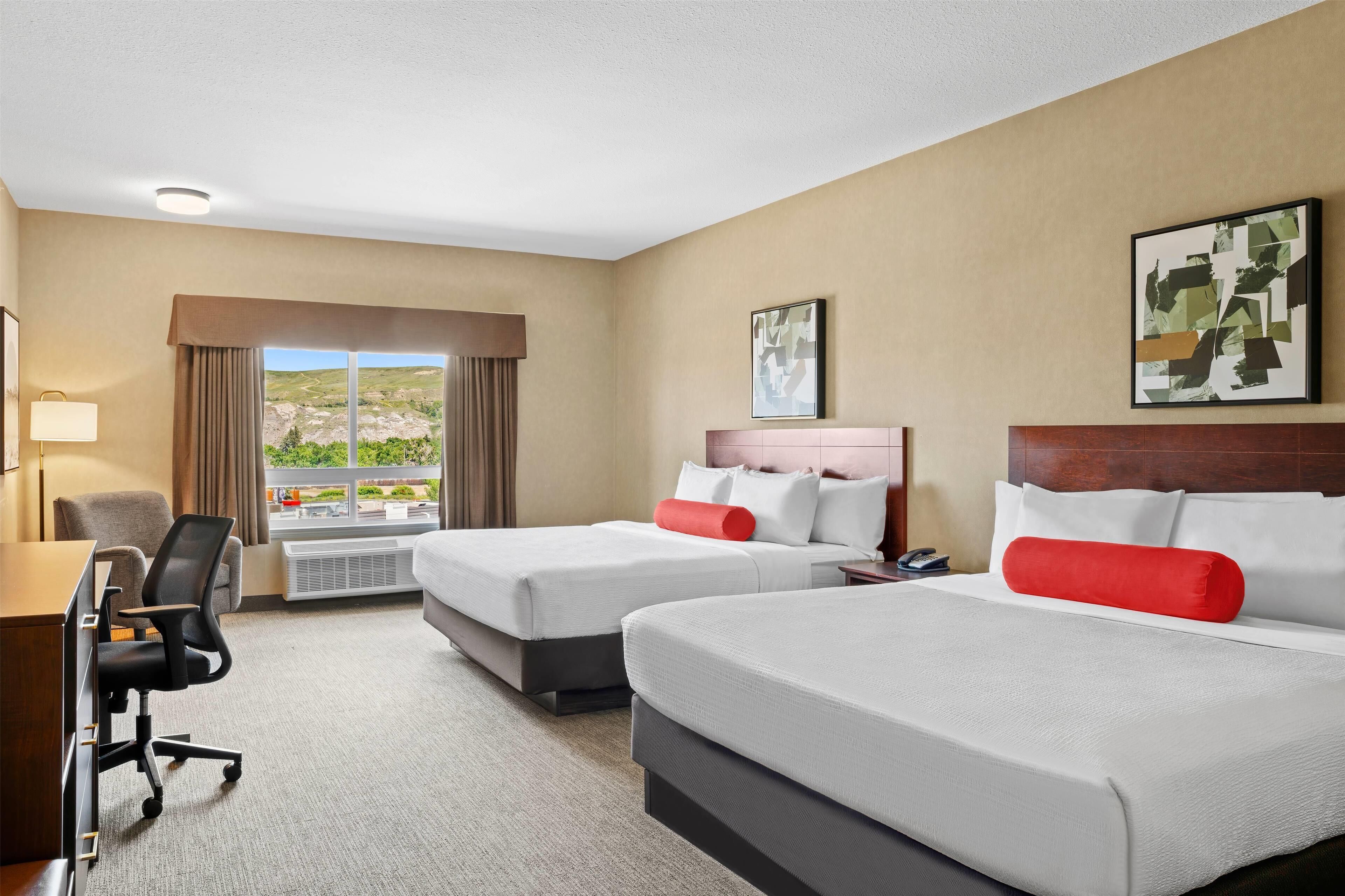 Une chambre d’hôtel avec deux lits, un bureau, une chaise et une grande fenêtre donnant sur un paysage verdoyant.
