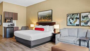 A hotel room with a bed, a sofa, a small table, a desk with a computer, a microwave, and a refrigerator.